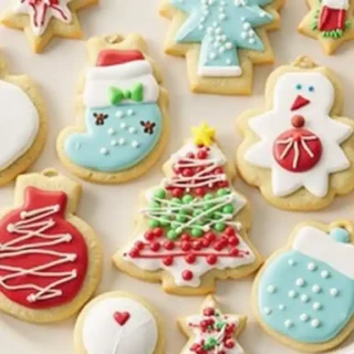 Festive Christmas ornament cookies decorated with vibrant red, green, and gold icing, placed on a rustic wooden table surrounded by pine branches. The background features warm twinkling fairy lights and a steaming mug of hot cocoa, creating a cozy and joyful holiday atmosphere."