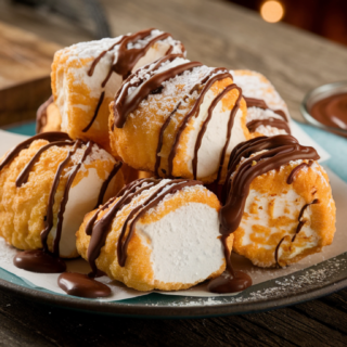 A beautiful plate of deep fried marshmallows arranged artfully, golden and crispy with a dusting of powdered sugar and a drizzle of chocolate sauce, served on a rustic wooden table. A small bowl of chocolate sauce and colorful sprinkles sit nearby, with a warm and inviting background.